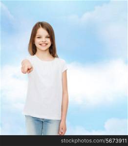 advertising, dream, childhood, gesture and people - smiling little girl in white t-shirt pointing finger on you over cloudy sky background