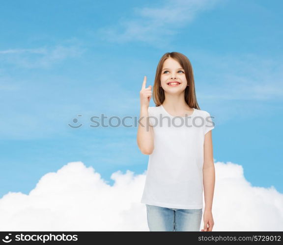 advertising, dream, childhood, gesture and people - smiling little girl in white blank t-shirt pointing finger up over blue sky background