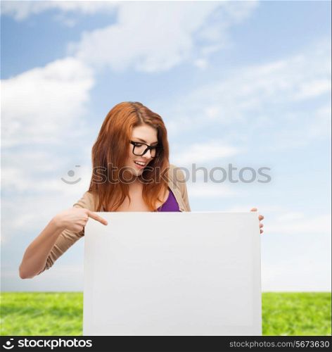 advertising, education and people concept - smiling teenage girl in glasses pointing finger to blank white board over blue sky and grass background