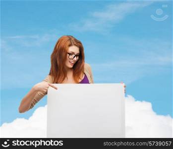 advertising, education and people concept - smiling teenage girl in glasses pointing finger to blank white board over blue sky and cloud background