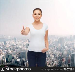 advertising, gesture and people concept - smiling young woman in blank white t-shirt showing thumbs up over city background