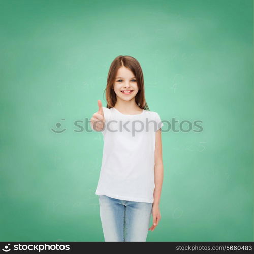 advertising, gesture, education, childhood and people - smiling little girl in white t-shirt showing thumbs up over green board background