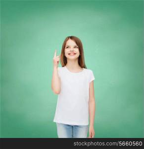 advertising, gesture, education, childhood and people - smiling little girl in white t-shirt pointing finger up over green board background