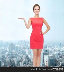 advertising, holidays and people concept - smiling young woman in red dress holding something on palm of her hand over city background