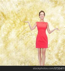 advertising, holidays and people concept - smiling young woman in red dress holding something on palm of her hands over yellow lights background