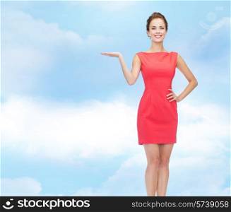 advertising, holidays and people concept - smiling young woman in red dress holding something on palm of her hand over blue cloudy sky background