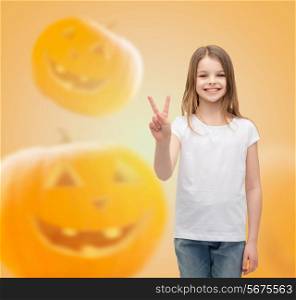 advertising, holidays, gesture and people concept - smiling little girl in white blank t-shirt showing victory sign over halloween pumpkins background