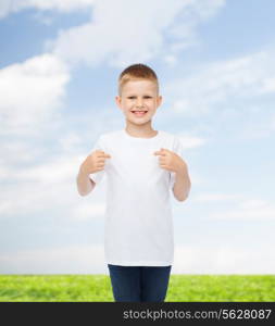 advertising, people and childhood concept - smiling little boy in white blank t-shirt pointing fingers at himself over natural background