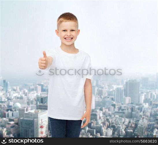 advertising, people and childhood concept - smiling little boy in white blank t-shirt showing thumbs up over city background
