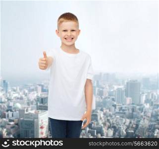 advertising, people and childhood concept - smiling little boy in white blank t-shirt showing thumbs up over city background