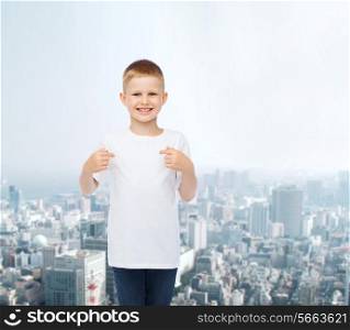 advertising, people and childhood concept - smiling little boy in white blank t-shirt pointing fingers at himself over city background
