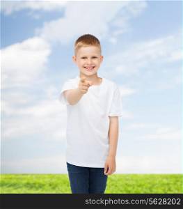 advertising, people, gesture and childhood concept - smiling boy in white blank t-shirt pointing finger at you over natural background