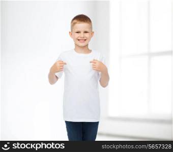 advertising, people, gesture and childhood concept - smiling boy in white blank t-shirt pointing finger himself over white room background