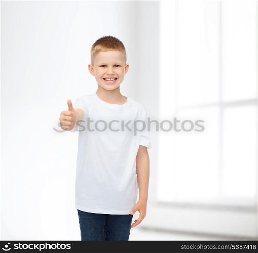 advertising, people, gesture and childhood concept - smiling little boy in white blank t-shirt showing thumbs up over white room background