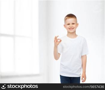 advertising, people, home and childhood concept - smiling little boy in white blank t-shirt making ok gesture over white room background