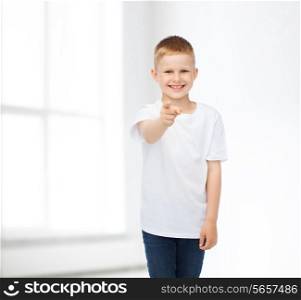 advertising, people, home, gesture and childhood concept - smiling little boy in white blank t-shirt pointing finger at you over white room background
