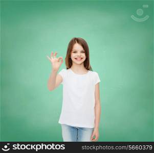advertising, school, education, childhood and people - smiling little girl in white t-shirt showing ok sign over green board background