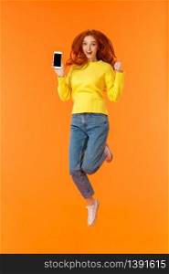 Advertising, technology and holidays concept. Vertical full-length shot cheerful gorgeous ginger girl, redhead woman jumping over orange background with smartphone, show thumb-up.. Advertising, technology and holidays concept. Vertical full-length shot cheerful gorgeous ginger girl, redhead woman jumping over orange background with smartphone, show thumb-up
