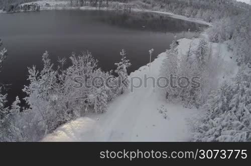 Aerial - car driving to the winter camp along snowy road by the lake and through pine forest with frozen trees