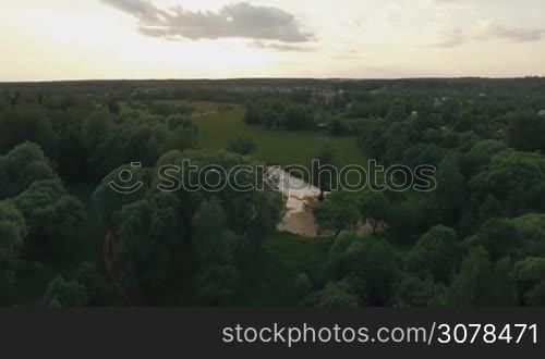 Aerial flight above the countryside in Russia. View of forest, small lake, green fields against blue sky with clouds in sunset time at summer. Beautiful landscape
