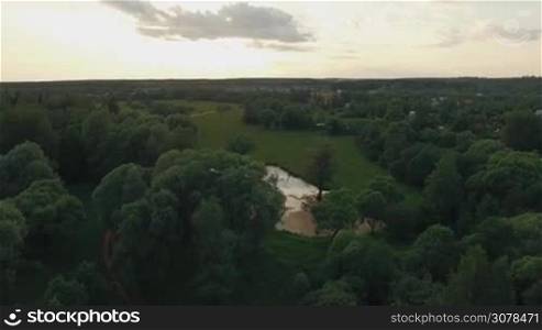 Aerial flight above the countryside in Russia. View of forest, small lake, green fields against blue sky with clouds in sunset time at summer. Beautiful landscape