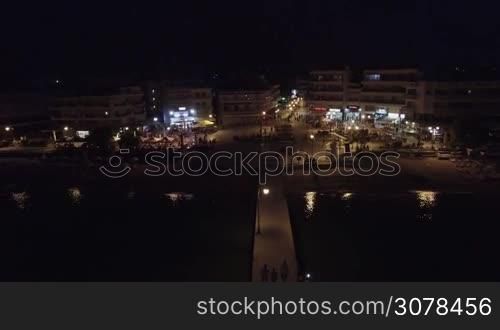 Aerial flight above the Mediterranean Sea, camera moving away from coast line with resort hotels along long pontoon at the sea at night, beach and pontoon, Thessaloniki, Greece