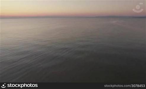 Aerial flight above the water of the sea against horizon line and clear sky at the sunset time