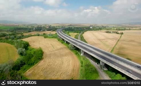 Aerial flight over the fields and next the highway. Aerial view with background of blue sky and clouds and mountains. On the fields are stored bales of hay next the highway