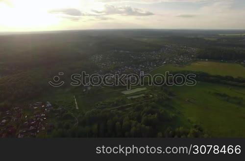 Aerial high level flight above the countryside in Russia. View of forest, country houses and agricultural fields against blue sky with clouds in sunset time at summer