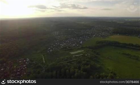 Aerial high level flight above the countryside in Russia. View of forest, country houses and agricultural fields against blue sky with clouds in sunset time at summer
