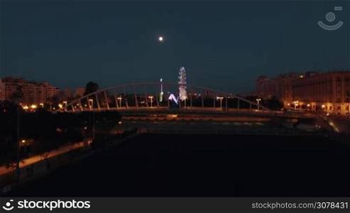 Aerial night view of lighted ferris wheel and bridge against sky with moon on the background, Valencia, Spain