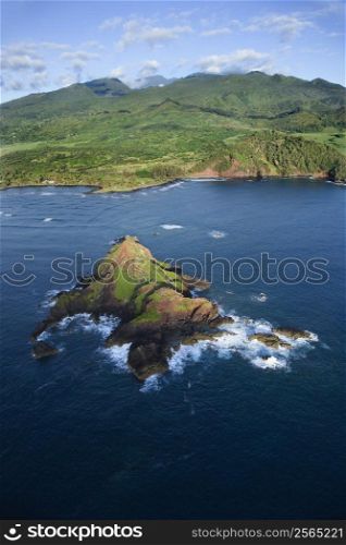 Aerial of rock jutting out of Pacific ocean off the coast of Maui, Hawaii with mountain landscape in background.