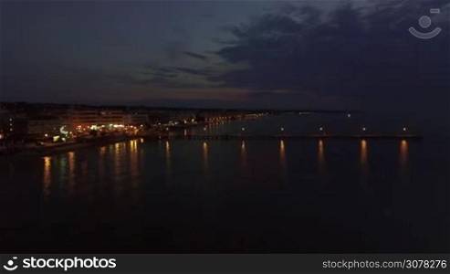 Aerial scene of resort at night. Quiet sea, pier with people and coastline illuminated with lanterns. Thessaloniki, Greece