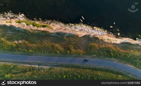 Aerial shot from above driving black car in a field by the rural road near a cliff at the seaside in summer at sunset