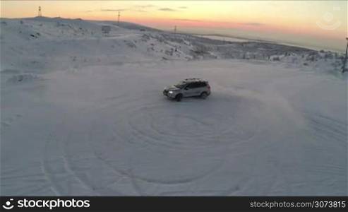 Aerial shot of a car drifting on snowfield making circles with view to the distant snow covered hills and evening sky. Beautiful northern nature