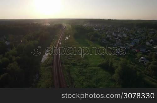 Aerial shot of a cargo train traveling through the green countryside with summer houses. Sunset scene, Russia