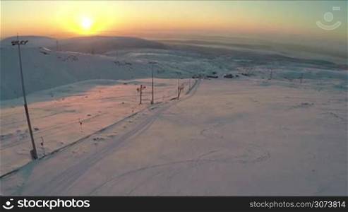 Aerial shot of distant ski resort and snowy hills with empty skiing run and ski-lift in foreground. Beautiful nature scene at sunset. Winter sports and recreation