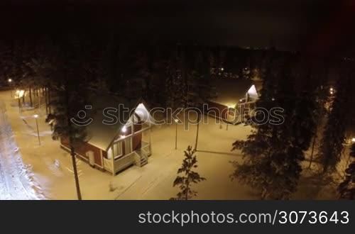 Aerial shot of guest country houses in winter pine wood at night. Territory with evergreen trees alight with lanterns. Winter holidays outside the town