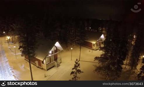 Aerial shot of guest country houses in winter pine wood at night. Territory with evergreen trees alight with lanterns. Winter holidays outside the town