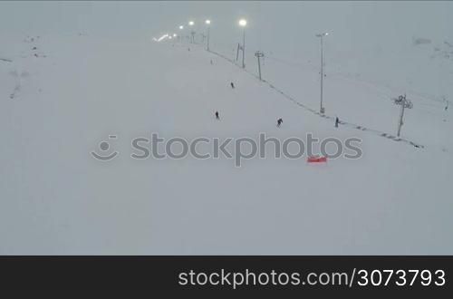 Aerial shot of people skiing and snowboarding down the slope with working ski lift and lampposts alongside. Winter sports and recreation
