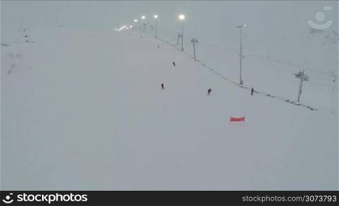 Aerial shot of people skiing and snowboarding down the slope with working ski lift and lampposts alongside. Winter sports and recreation