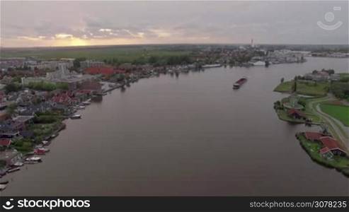Aerial shot of ship sailing along the river flowing through the township in Netherlands. View at sunset