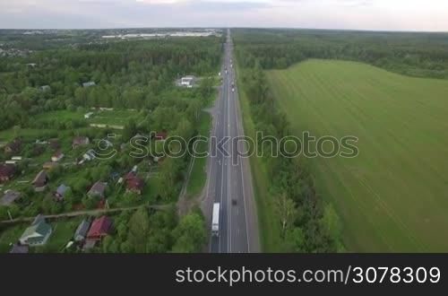 Aerial shot of the road with transport traffic running along the village, woods and fields. Russian countryside