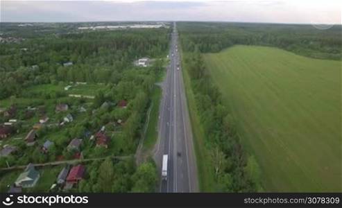 Aerial shot of the road with transport traffic running along the village, woods and fields. Russian countryside
