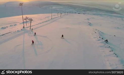 Aerial shot of two skiers and two snowboarders moving down the ski-run at sunset. Snowfields and hills in the distance. Doing winter sports