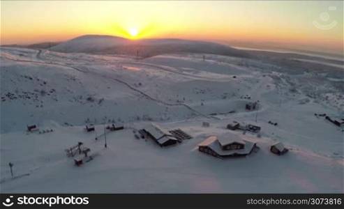 Aerial shot of winter ski resort. Snowy area with wooden buildings, golden sun going down behind the mountains. Khibins, Russia