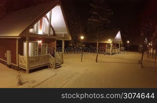 Aerial - small wooden guest houses in winter forest. Quiet evening, snow in warm lights of lanterns