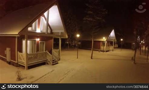 Aerial - small wooden guest houses in winter forest. Quiet evening, snow in warm lights of lanterns