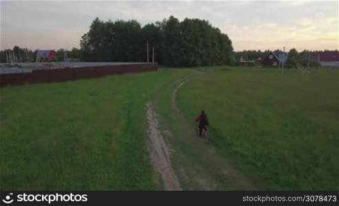 Aerial steadicam back view of boy riding a bike along countryside road between agricultural fields to the houses at summer, Russia