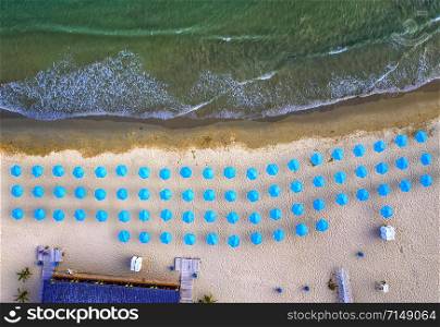 Aerial top view of a beautiful small beach with blue umbrellas, and sea waves.